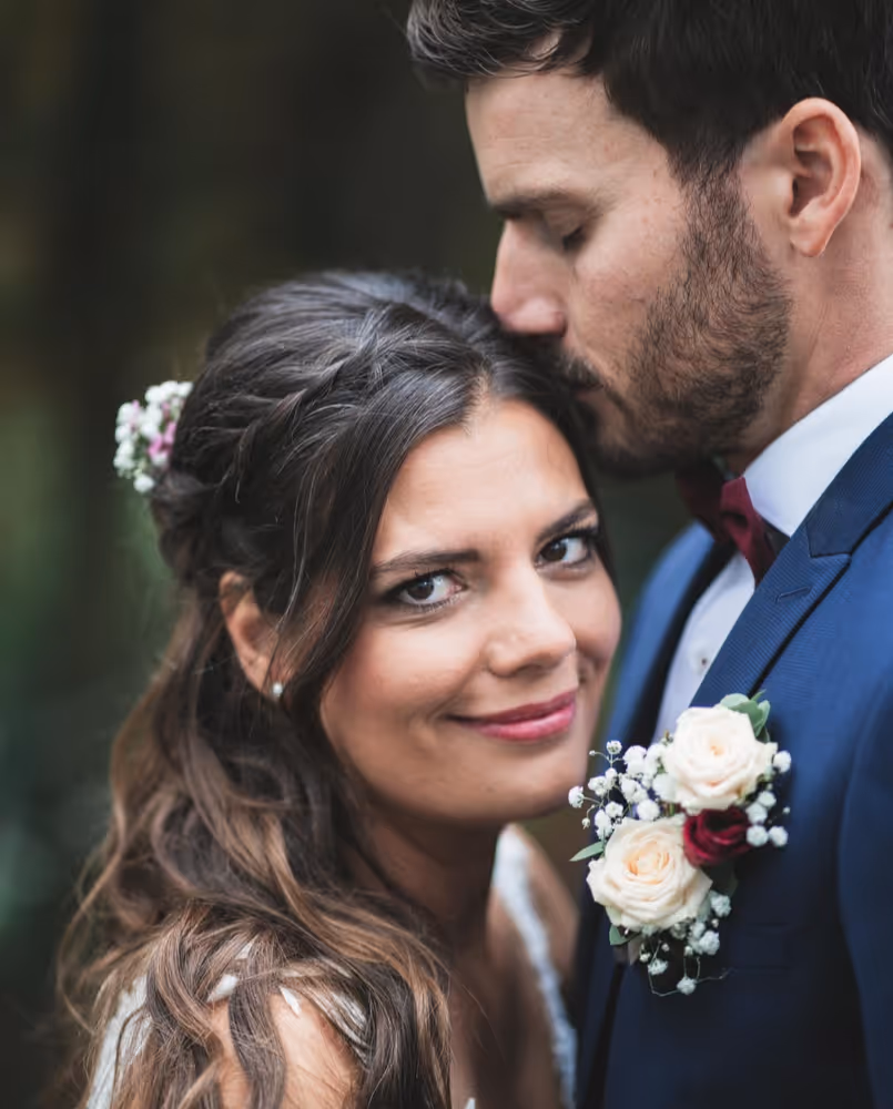 Glückliche Braut blickt direkt in die Kamera, während der Bräutigam ihr zärtlich die Stirn küsst. Portrait des Brautpaares im Freien mit Fokus auf Blumen-Details und Emotion.