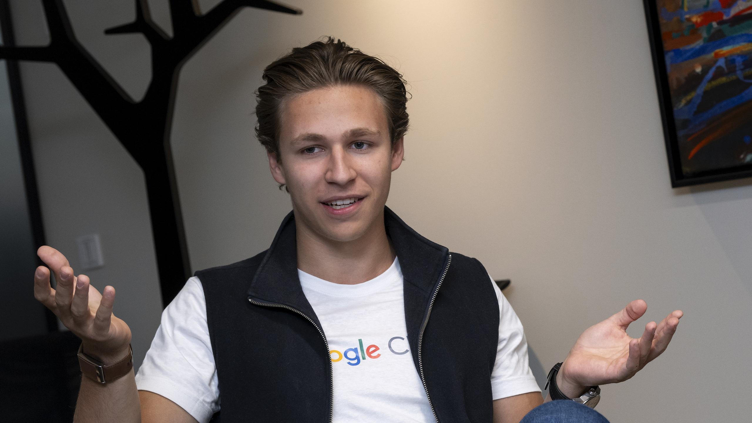 William Lindholm, Ung mann med bakoverstrøket hår som har på seg en hvit Google-T-skjorte og svart vest, og som gestikulerer med begge hender mens han sitter innendørs.