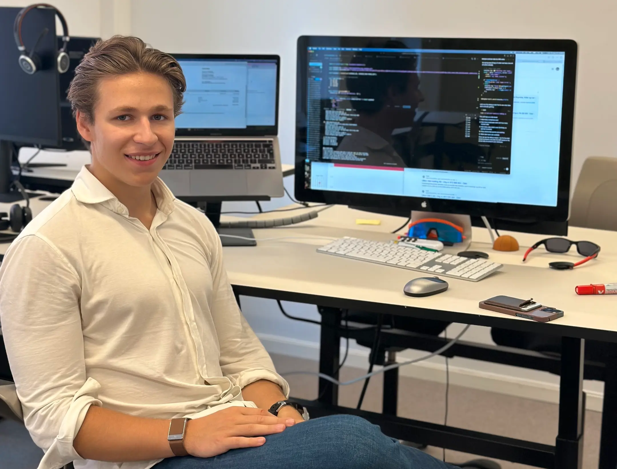 William Lindholm, Ung mann i hvit skjorte som sitter i en kontorstol foran et skrivebord med to dataskjermer som viser koding og dokumenter.