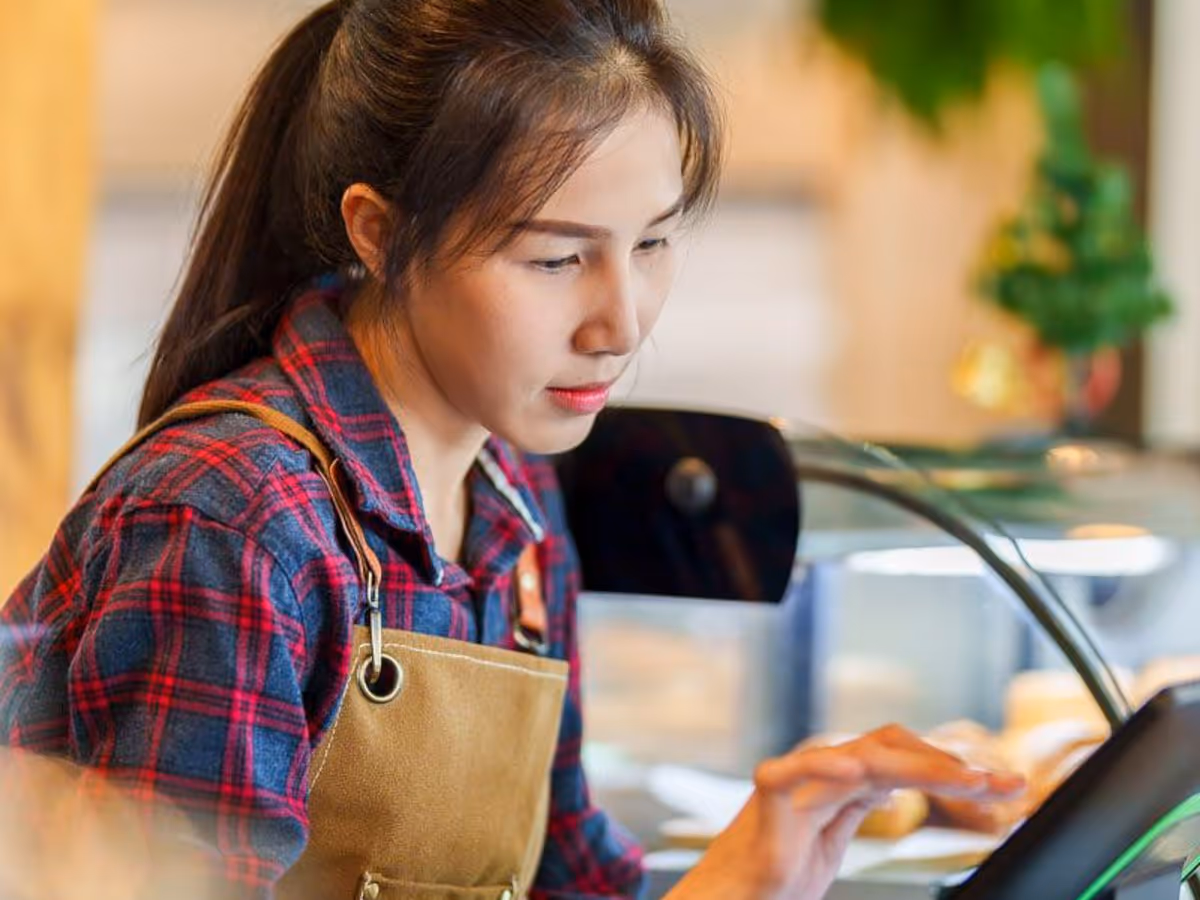 Een Aziatische vrouw in een geruit overhemd en een bruin schort bedient een restaurant met touchscreenbediening.