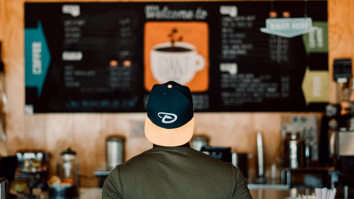 Man met een baseballpet leest het menu van een koffiebar, met kleurrijke inspiratie voor kopjesontwerpen ernaast.