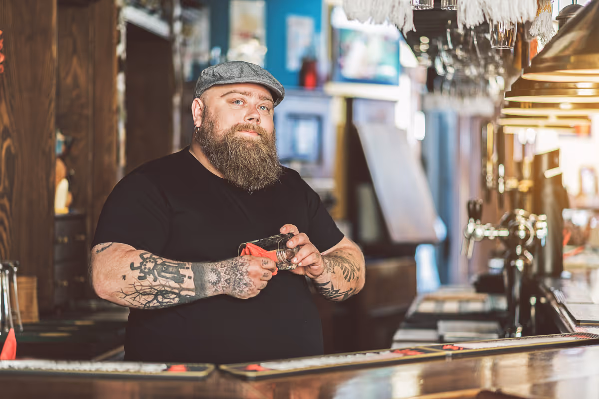 Een getatoeëerde barman poetst vakkundig glaswerk aan een houten toonbank in een trendy baromgeving.
