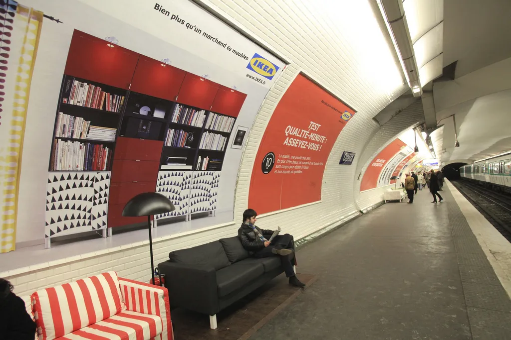 IKEA advertising takeover in a Paris metro station, with IKEA sofas placed on the platform and furnished room scenes printed along the tiled wall.