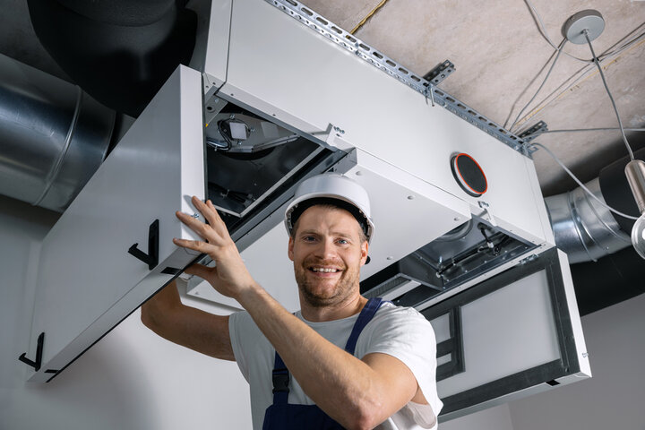 Smiling technician installing HVAC unit on ceiling in industrial setting