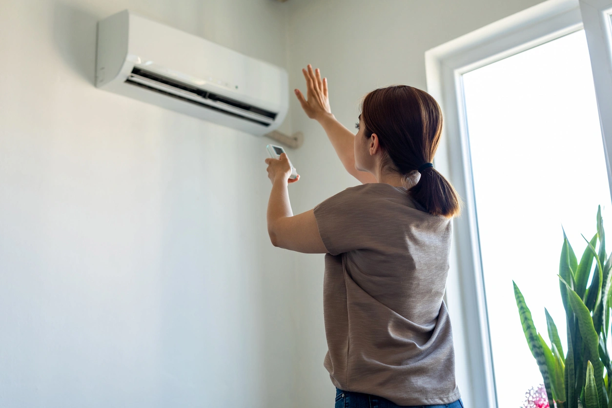 A woman adjusts an air conditioner with a remote control in a bright room.