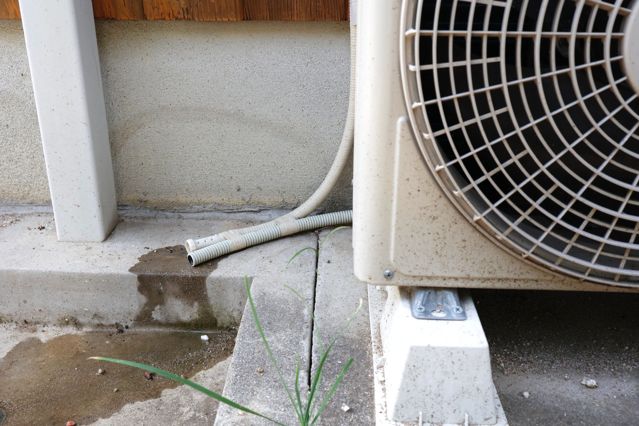 Close-up of an air conditioner's outdoor unit with a visible drainage pipe dripping water onto the ground. 