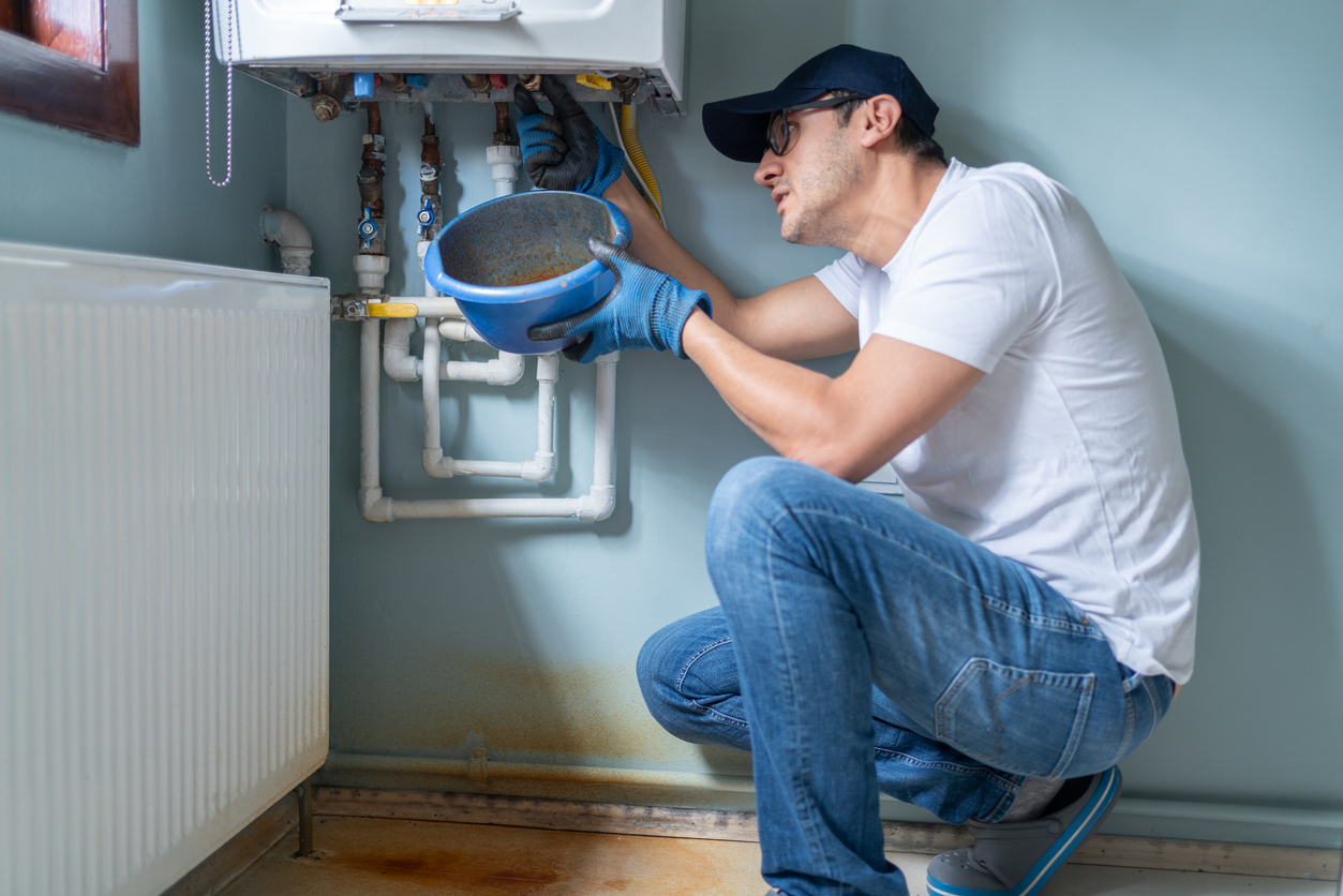 A man in a white shirt, cap, and jeans kneels by pipes, examining a boiler while holding a basin. 