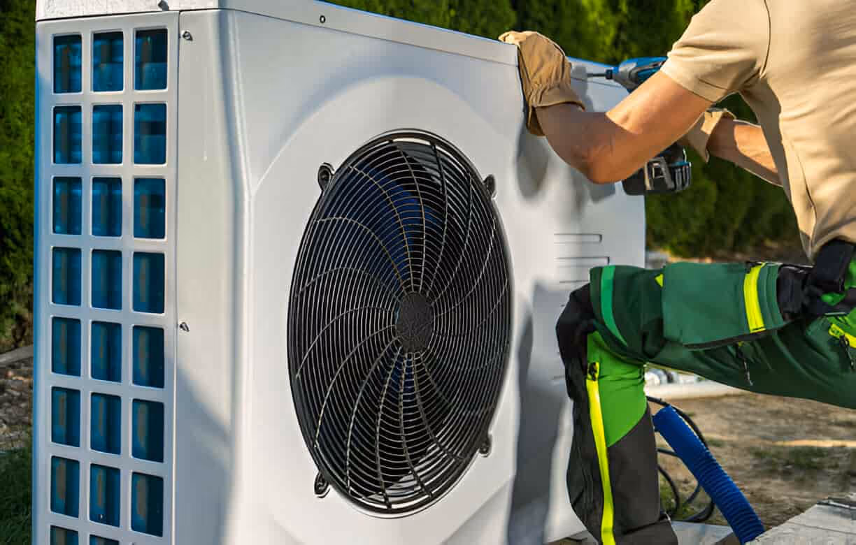 Technician servicing an outdoor heat pump with large fan and blue grid