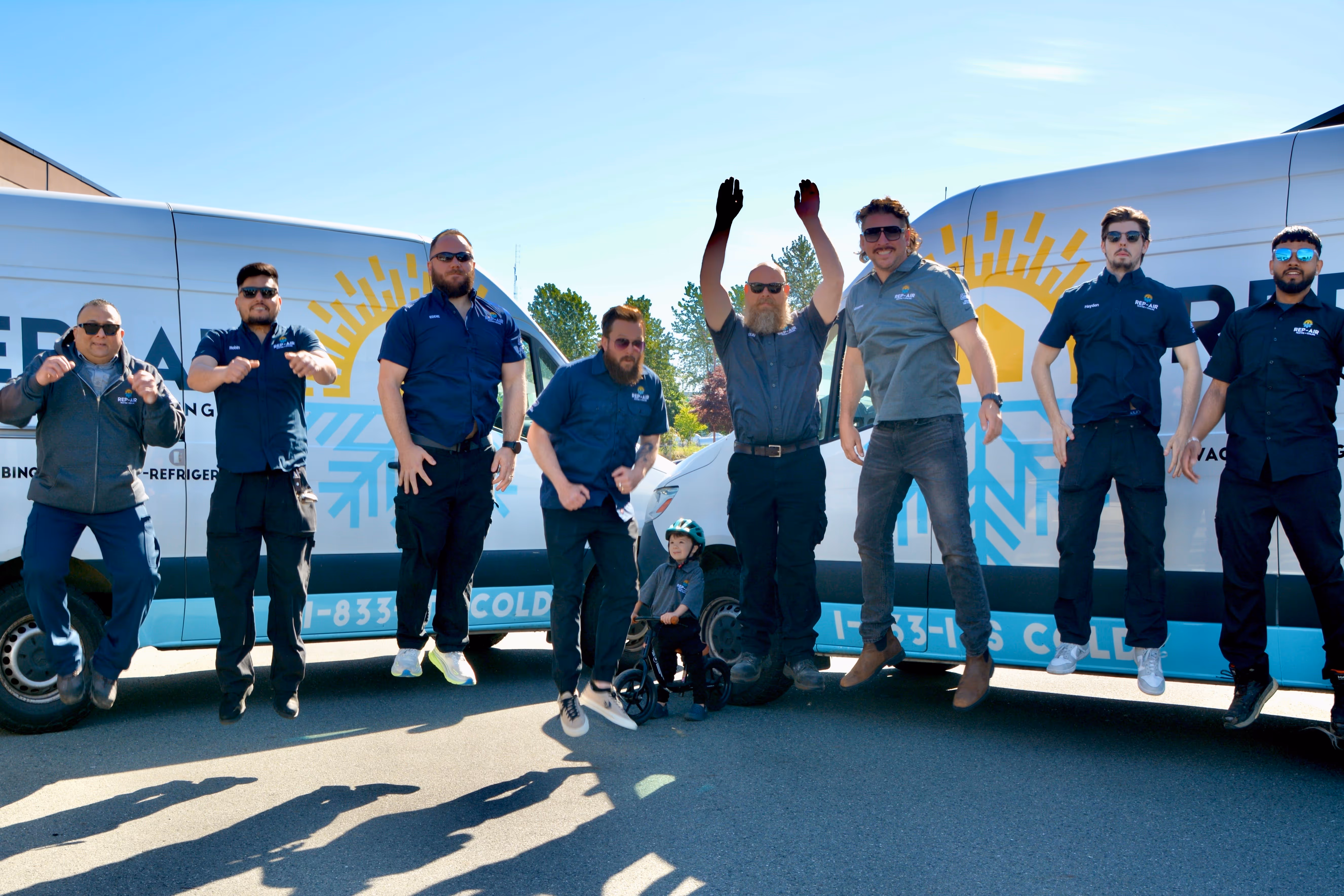 Group of nine men and one child jumping in front of two white service vans with company logos under a clear blue sky.