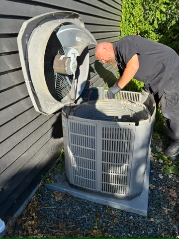 Man in black clothing inspecting an open outdoor air conditioning unit beside a dark wooden wall.