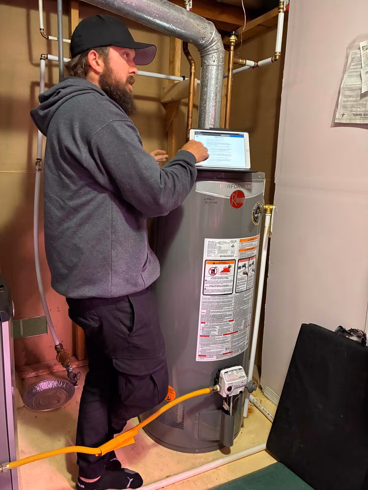 Man in a gray hoodie and black cap interacting with a tablet on top of a gray Rheem water heater in a utility room.