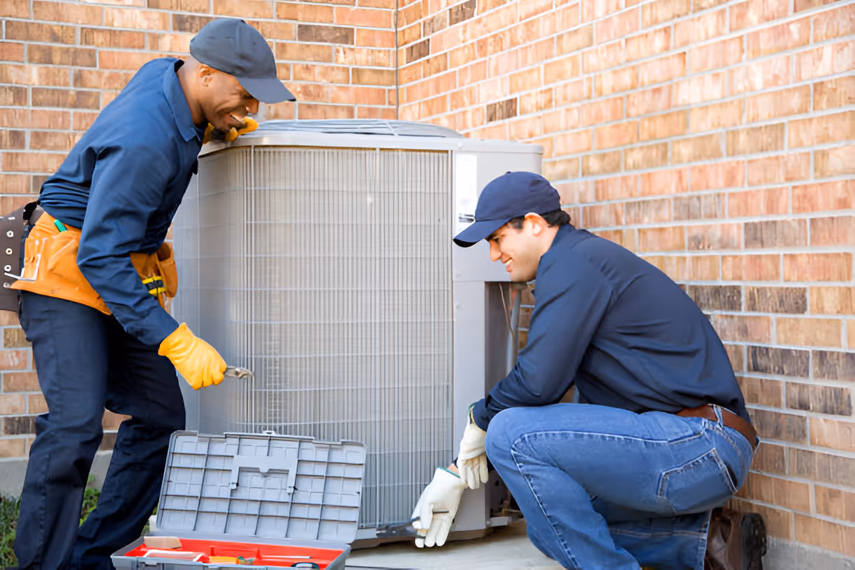 Two technicians repairing an air conditioning unit outside a brick building