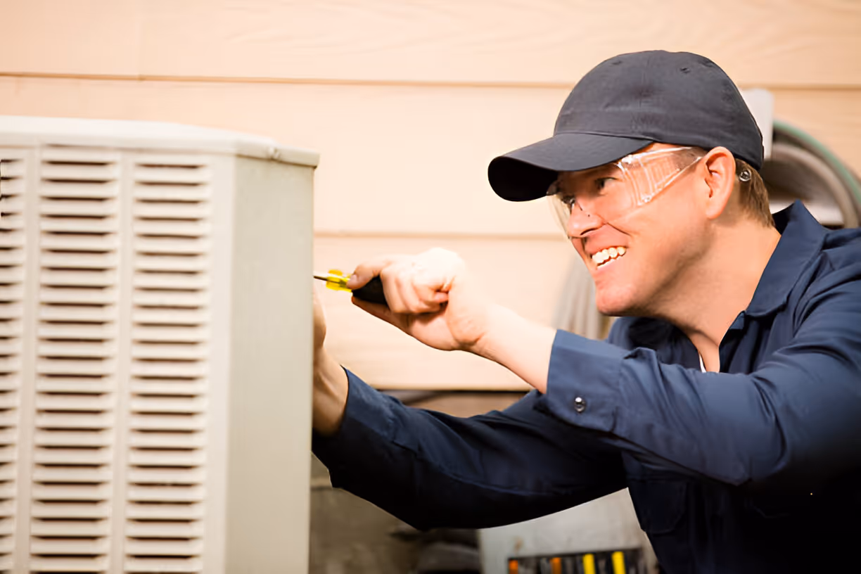 Technician in safety glasses and cap repairs HVAC unit with screwdriver