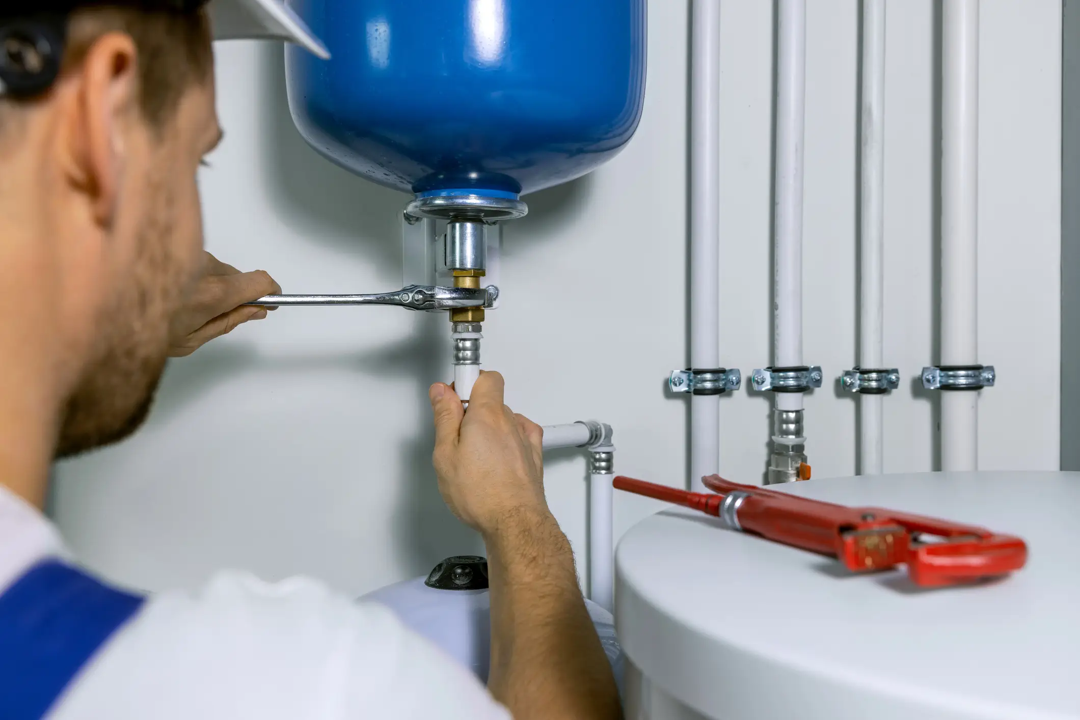A plumber in a white shirt and blue overalls uses a wrench to tighten a fitting on a blue expansion tank. White pipes and a red pipe wrench are also visible.