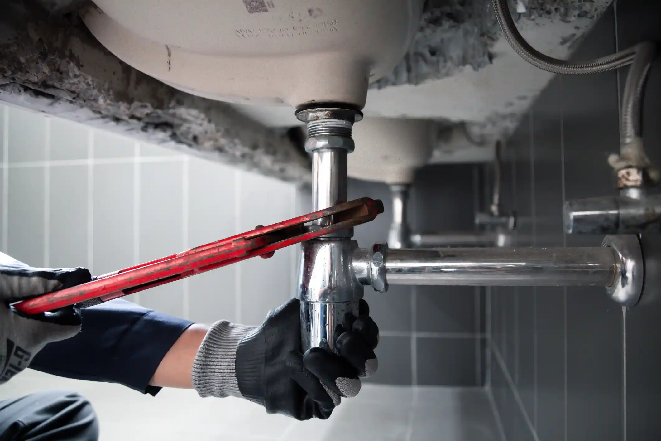 A person wearing work gloves uses a red pipe wrench to tighten a chrome drain assembly under a white sink in a commercial restroom with grey tiles.