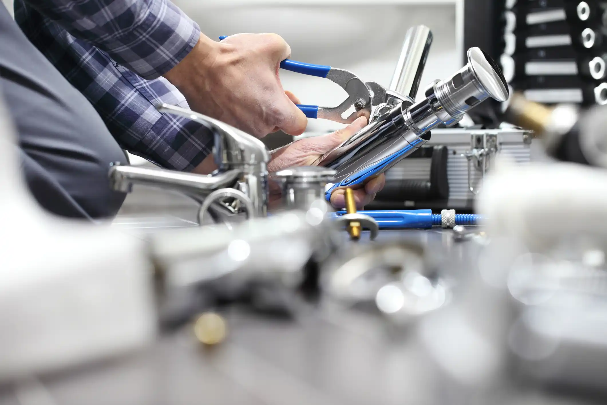 A plumber in a plaid shirt uses blue-handled pliers to assemble a chrome faucet component on a workbench. Various plumbing fixtures and a silver tool case are visible in the blurred foreground and background.