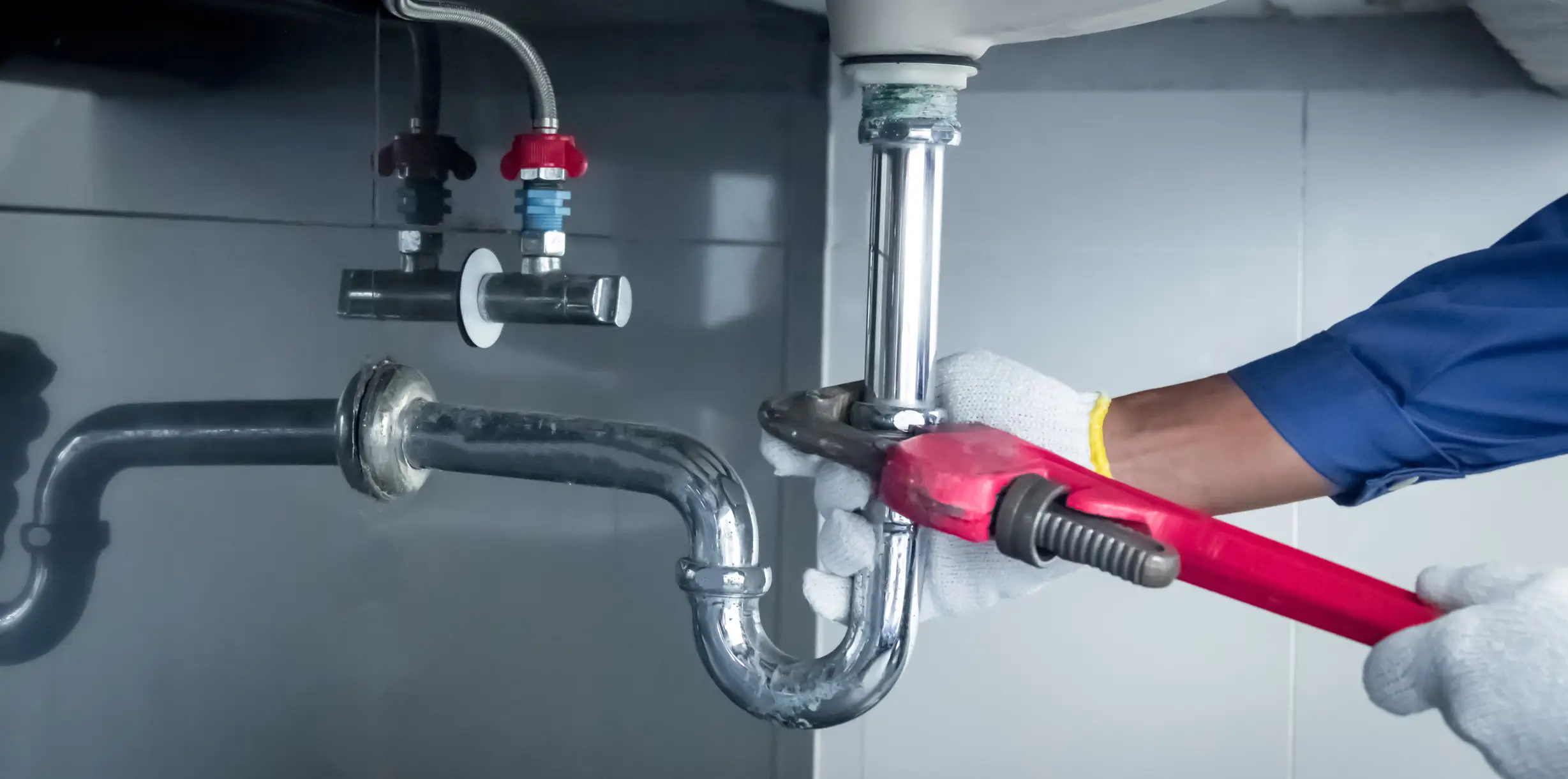 A plumber in a blue uniform and white gloves uses a red pipe wrench to tighten a chrome P-trap under a sink. Shut-off valves with red and blue handles are visible on the tiled wall.