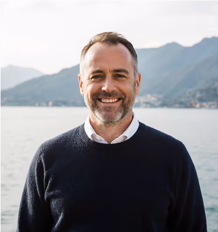 Portrait d’un client souriant devant le lac Léman, portant un pull marine élégant. Représente un entrepreneur satisfait après un projet digital réussi.