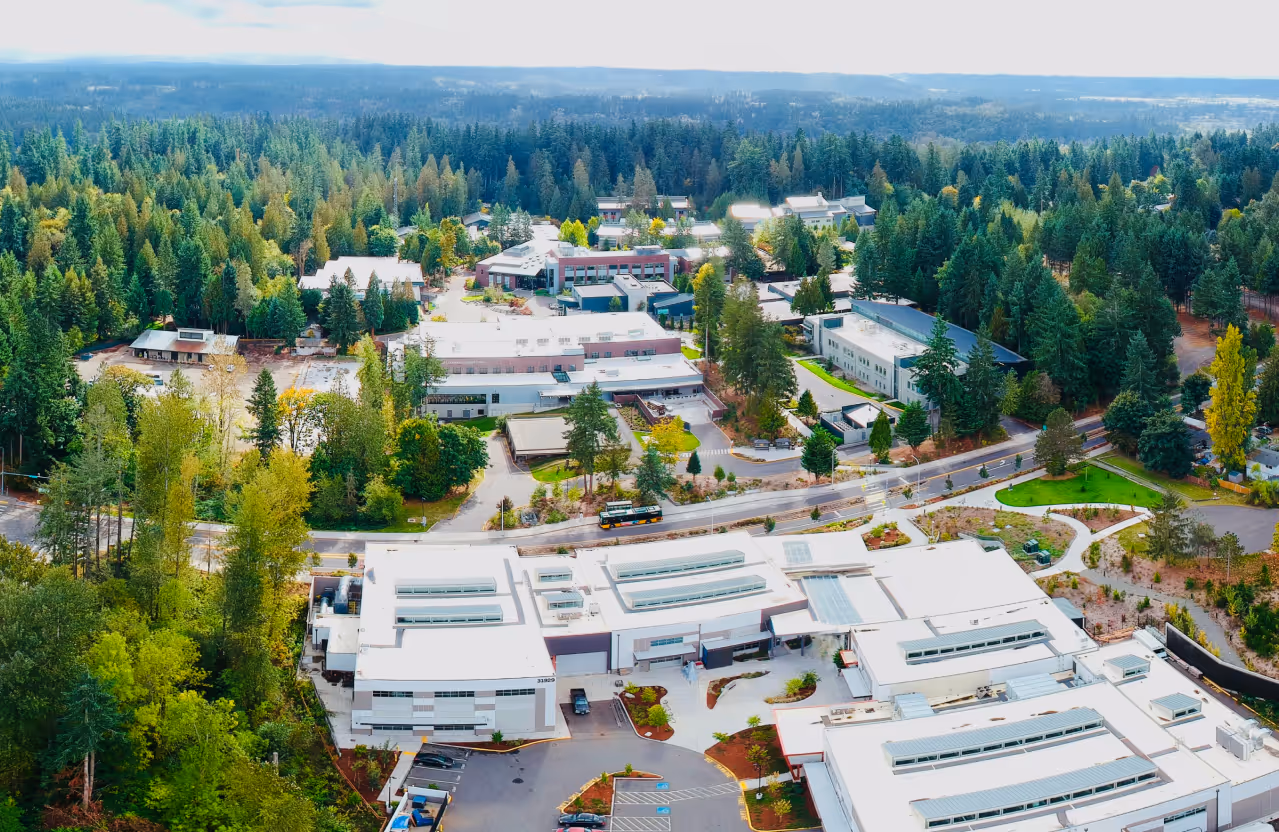 Aerial view of a large campus surrounded by dense green forest with multiple modern buildings and parking areas.