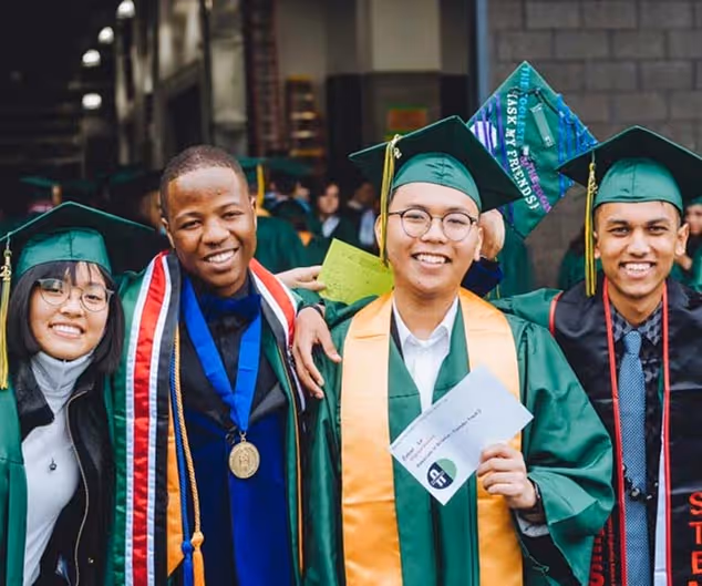 Four diverse graduates in green caps and gowns smiling and posing together outdoors.