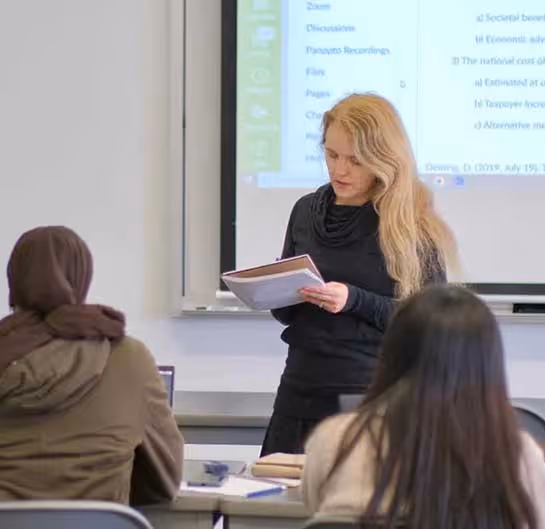 Teacher with long blonde hair reading from papers in front of a classroom with two seated students.