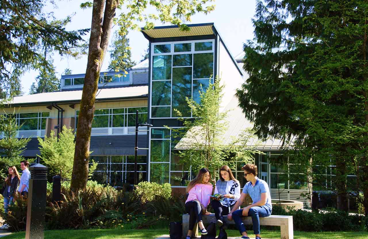 Three students sitting on a bench outside a modern campus building surrounded by trees and greenery, with two students walking in the background.