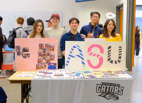 Five young adults standing behind a table with posters spelling 'ASU' and Green River Gators banner in a bright indoor setting.