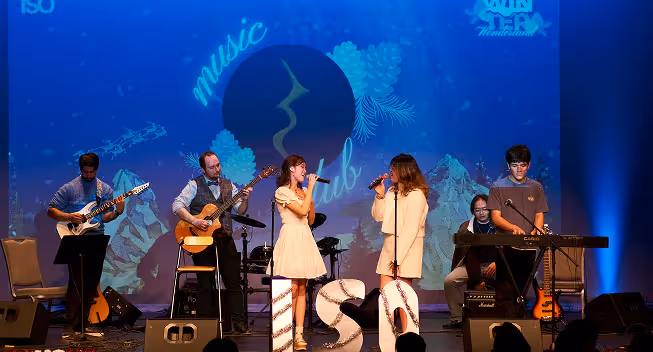 Band with two female singers in white dresses performing on stage with guitarists, keyboardist, and drummer under blue lighting.