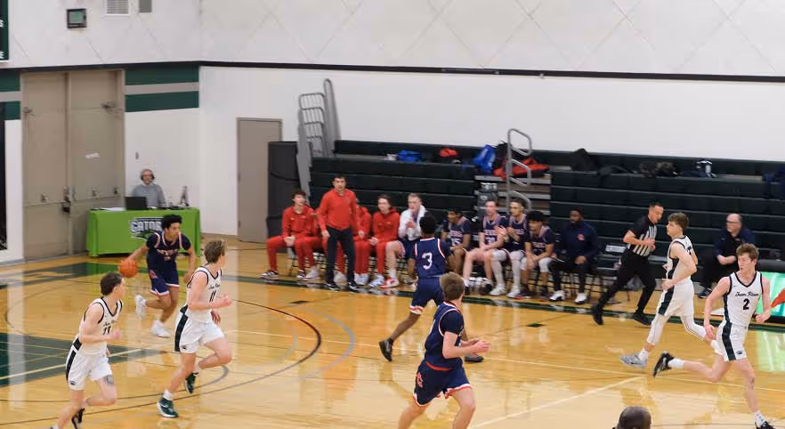 High school basketball game in progress with players from two teams running on a gym court and coaches on the sidelines.