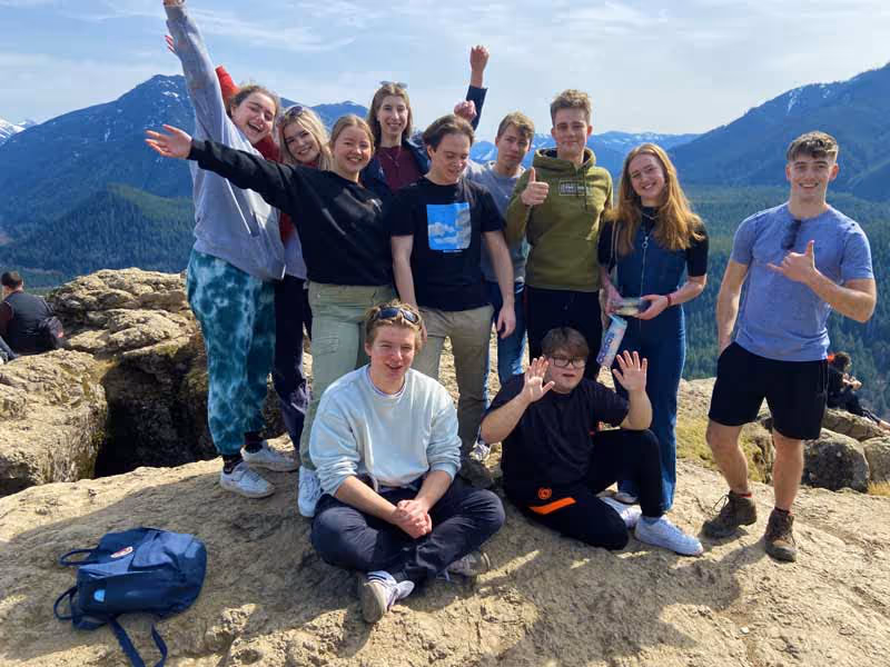 Group of young people posing and smiling on a rocky outcrop with forested mountains in the background.