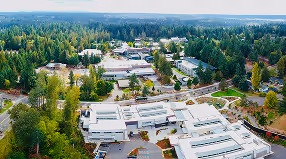 Aerial view of a modern school campus with multiple white-roofed buildings surrounded by dense green trees and forest.