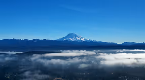 Snow-capped mountain peak under a clear blue sky above a layer of clouds and dark hills.