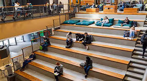 Modern auditorium with tiered wooden seating and people sitting and talking.