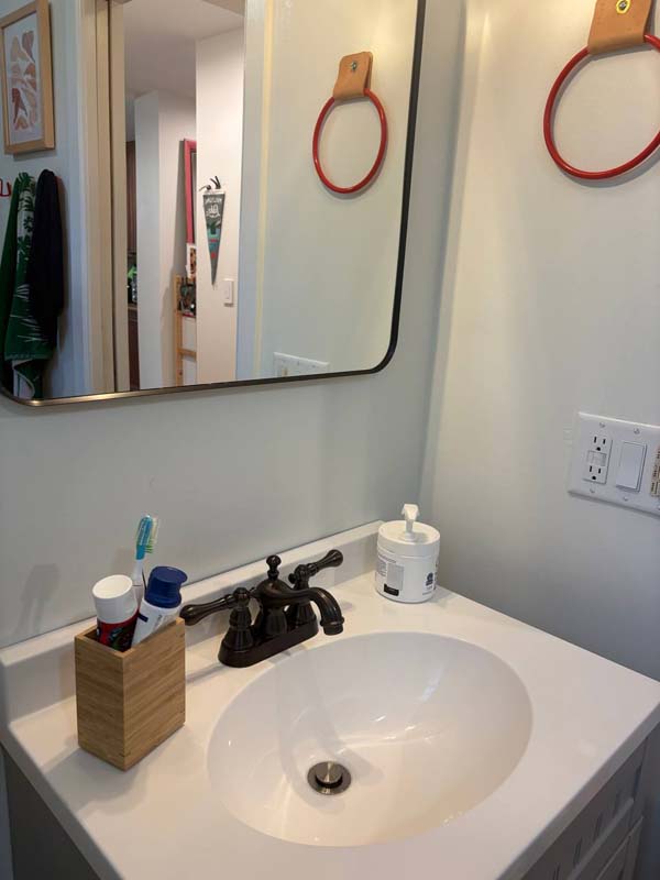 White bathroom sink with black faucet, toothbrush holder with toothbrush and toothpaste, dispenser container, and two red towel rings on a white wall.