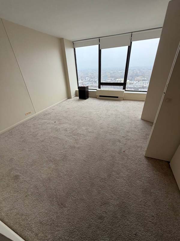 Empty beige carpeted room with large windows showing a cityscape view and a small black side table near the window.