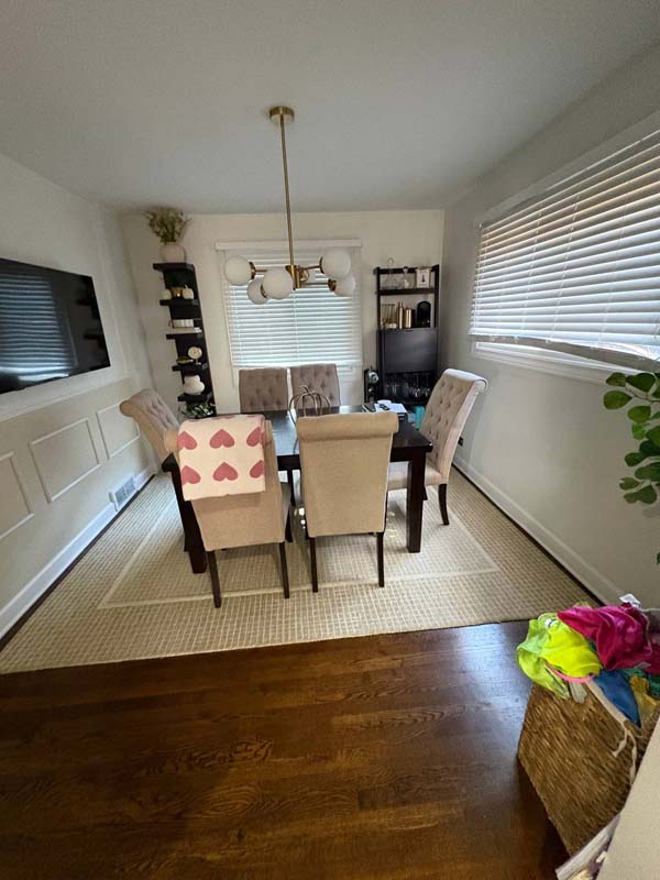 Dining room with a dark wooden table, six beige upholstered chairs, white pendant light fixture, two tall shelving units, and windows with blinds.