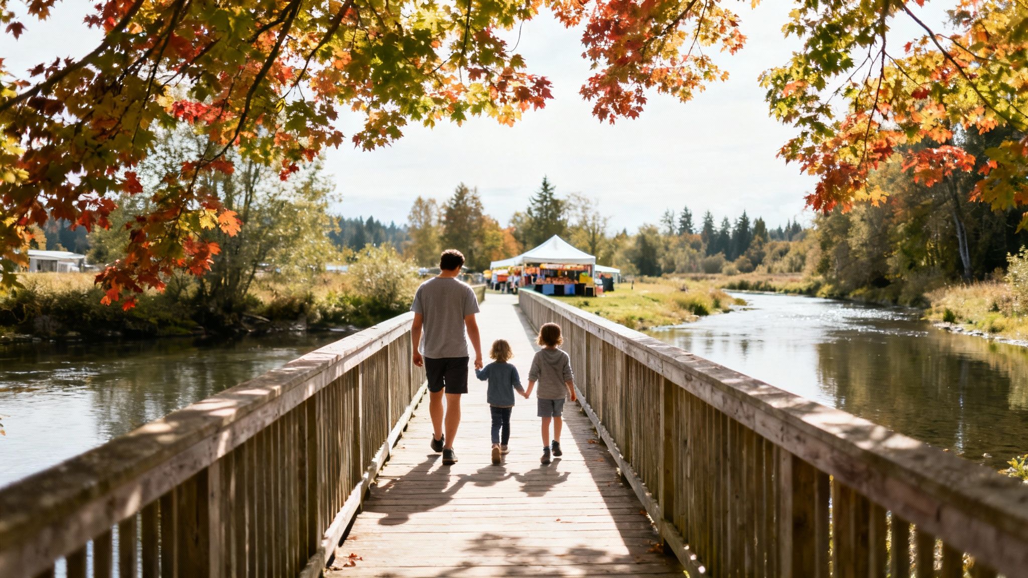 Father and two children walk on a wooden bridge over a river towards an outdoor market, framed by autumn leaves.