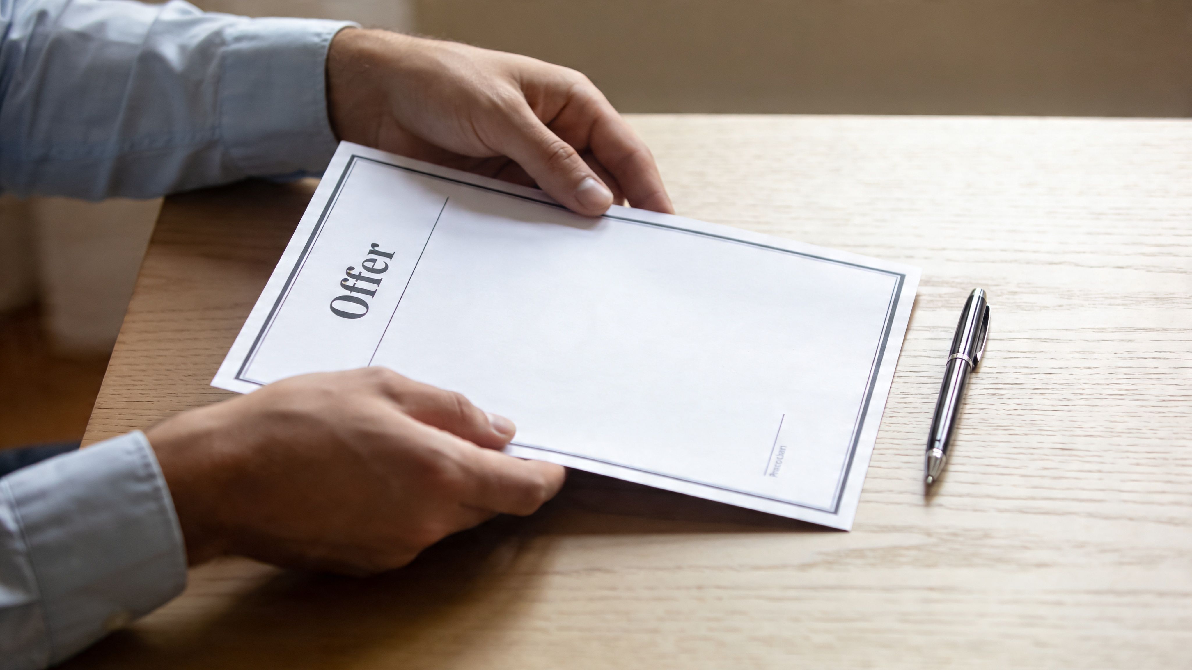 A professional man holding a formal job offer document on a wooden table with a pen.