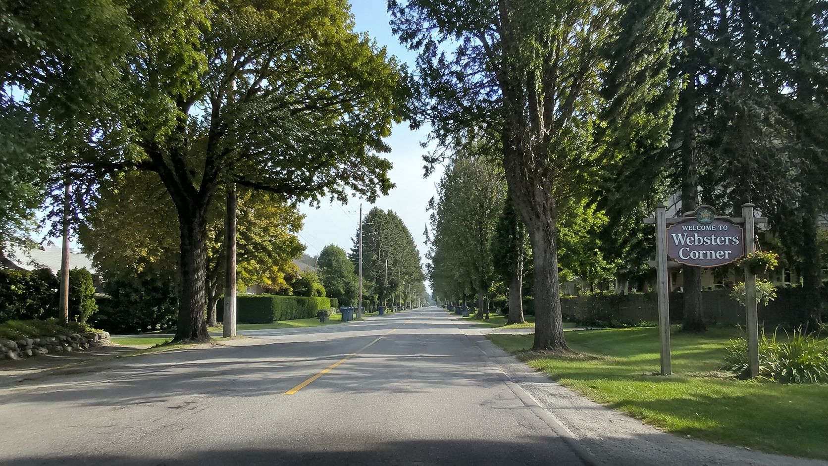 A quiet tree-lined road leading into the community of Websters Corner with a welcome sign on display.