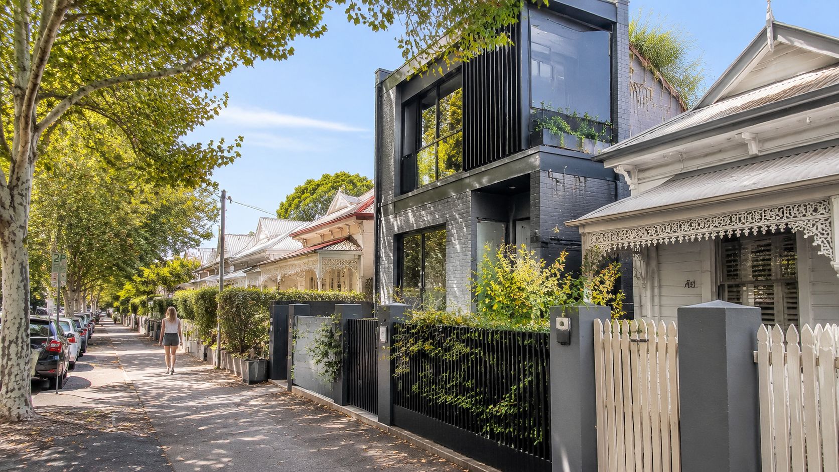 A modern black architectural home situated between traditional period houses on a sunny tree lined street.
