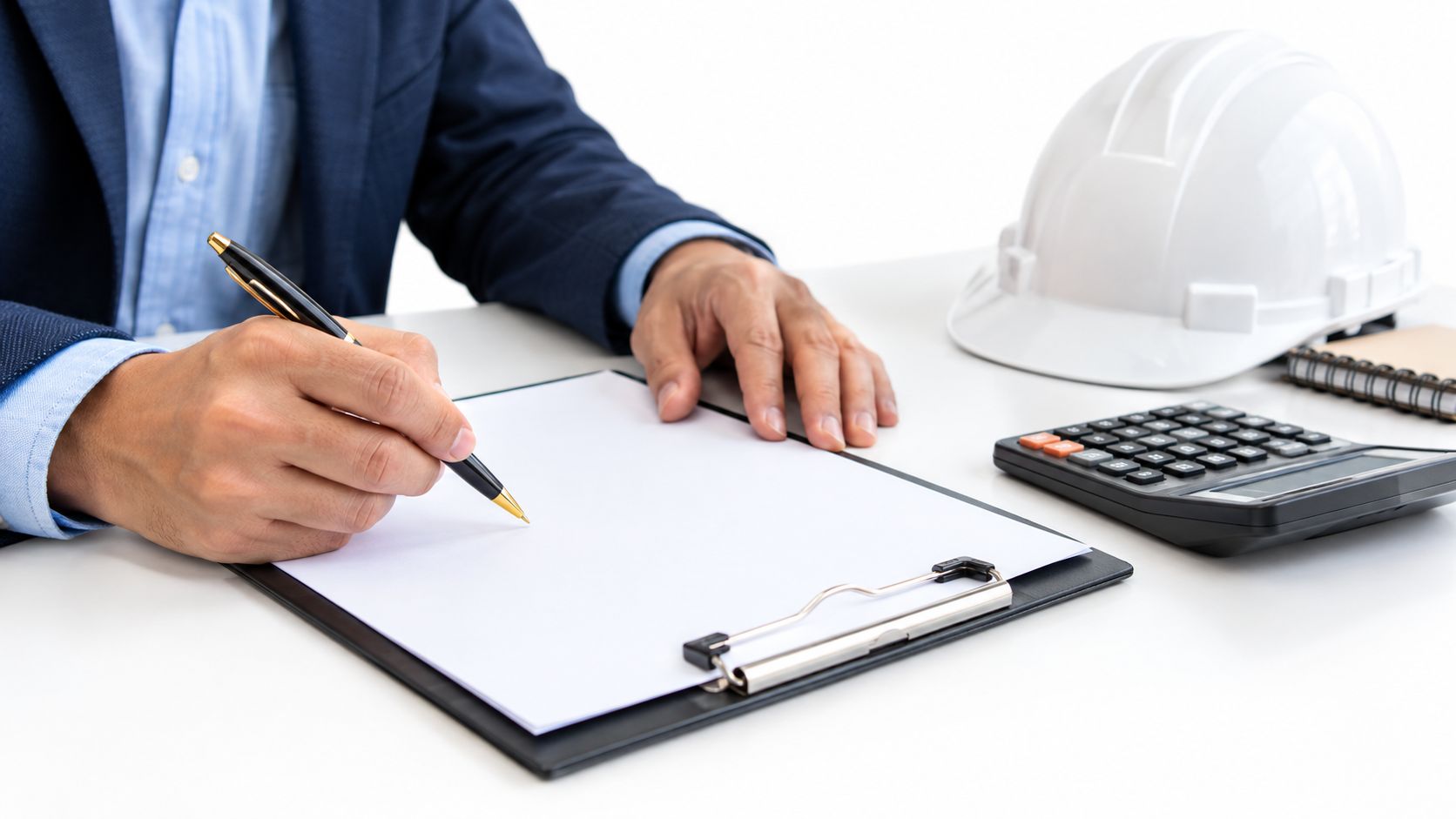 A professional in a business suit signing documents on a clipboard near a calculator and hard hat.