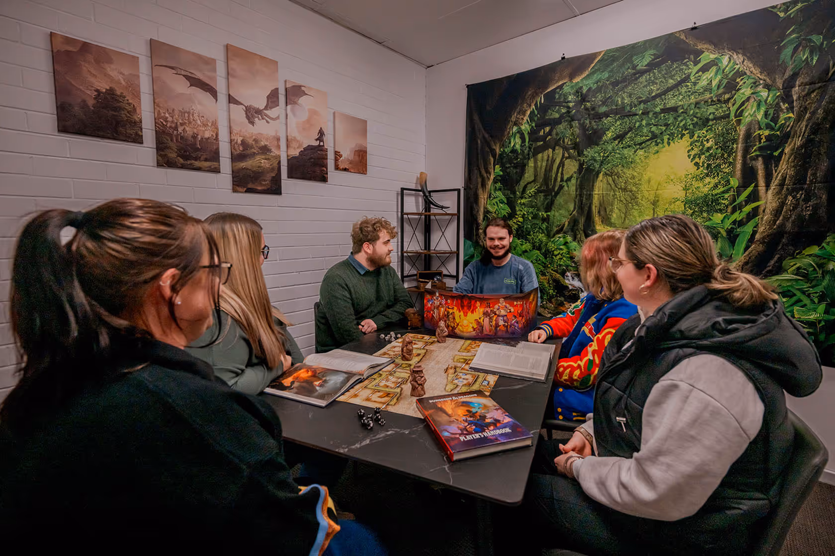 Group of six people gathered around a table playing a tabletop role-playing game with dice, character books, and a game master screen in a room with fantasy-themed wall art.