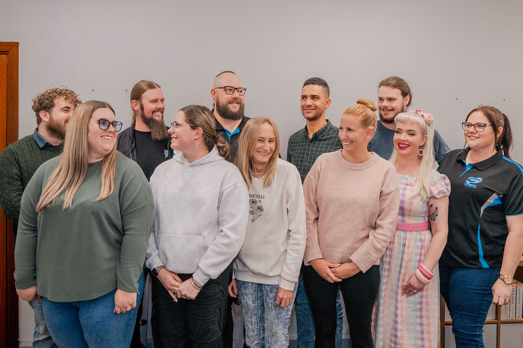 Group of eleven smiling people standing closely together indoors against a plain wall.