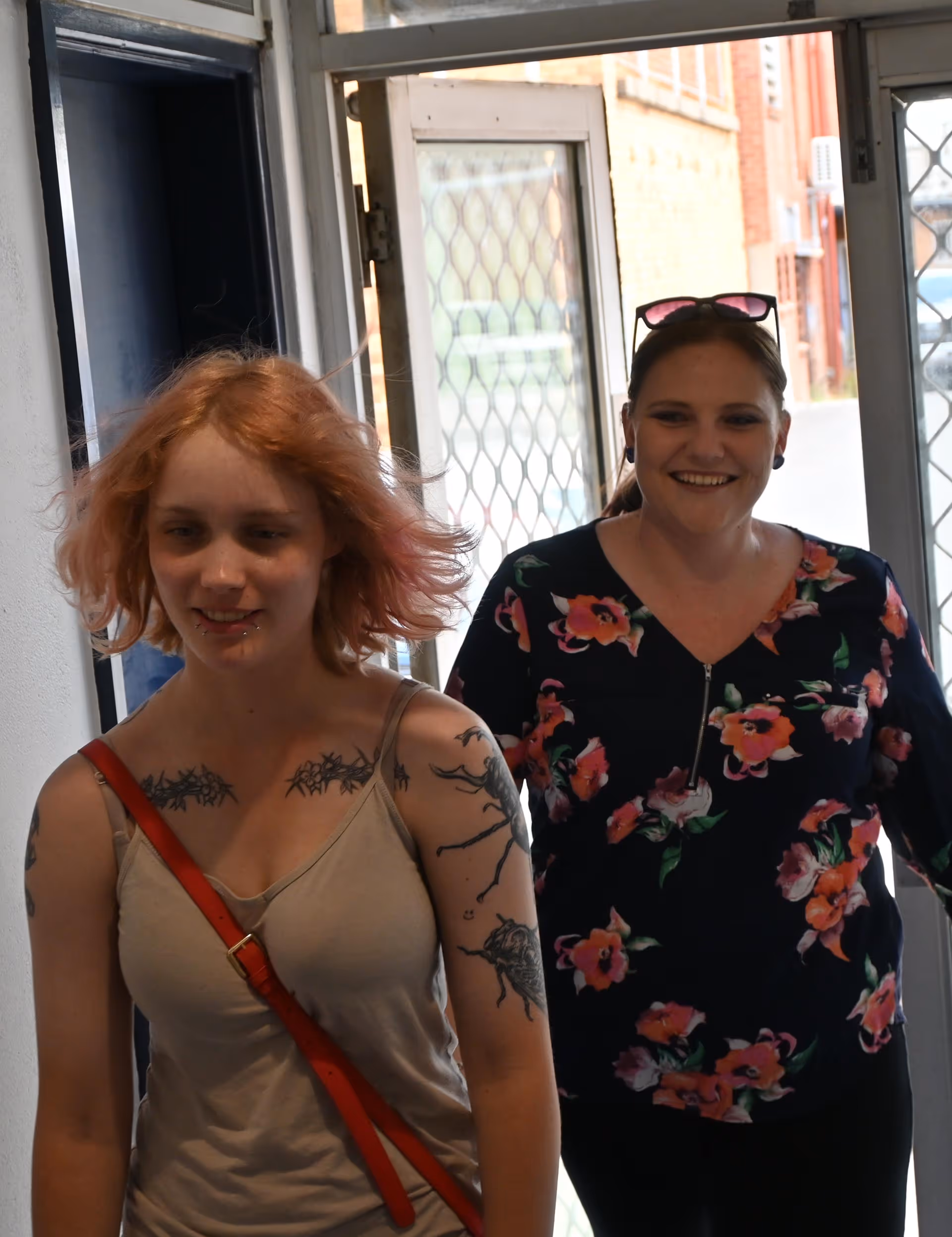 Two women entering a building, one with pink hair, tattoos, and a red strap across her shoulder, the other smiling and wearing a floral blouse with sunglasses on her head.