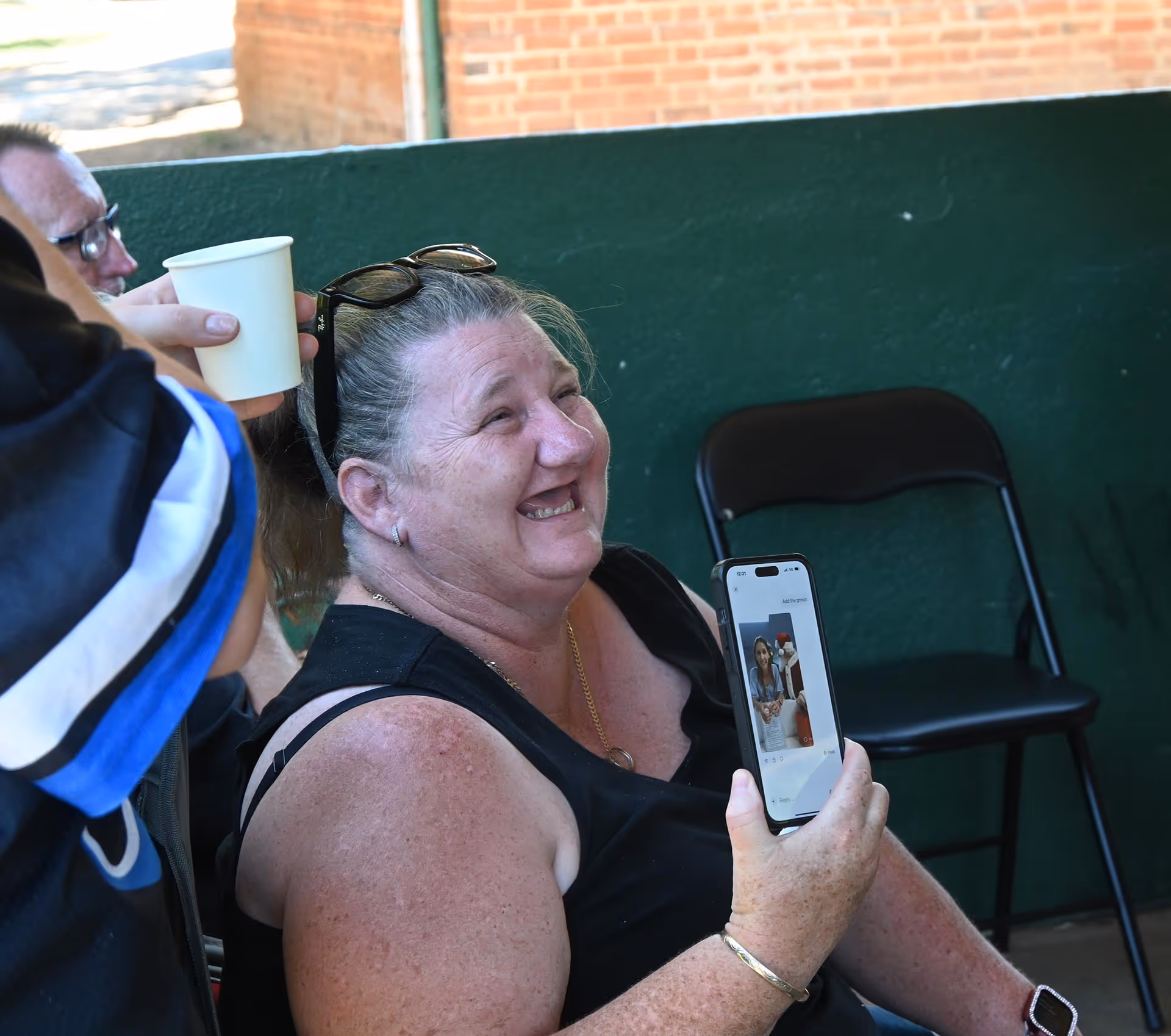 Smiling woman holding up a smartphone displaying a photo while sitting next to a person holding a white paper cup.