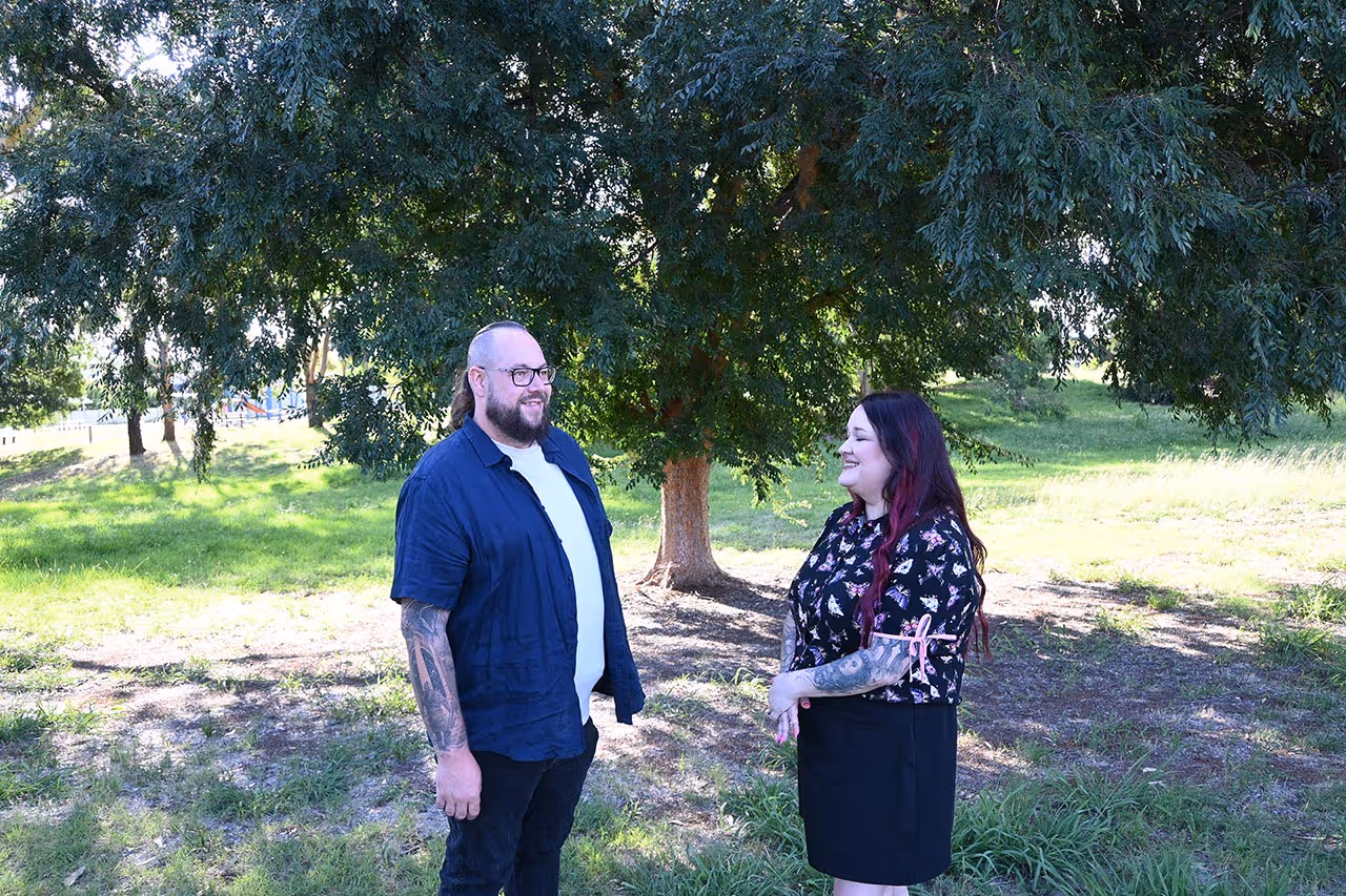 A man and a woman with tattoos standing and smiling at each other under a large tree in a grassy park.