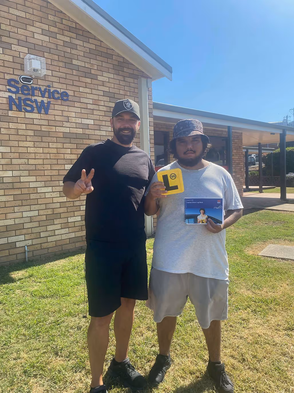 Two men standing on grass outside a Service NSW building, one holding a yellow learner driver plate and a learner driver logbook.