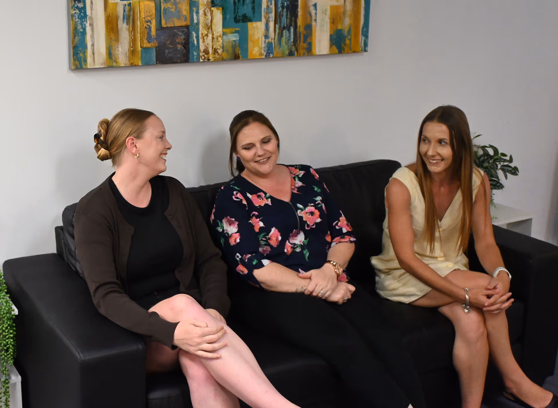 Three women sitting on a black couch smiling and engaged in conversation indoors.