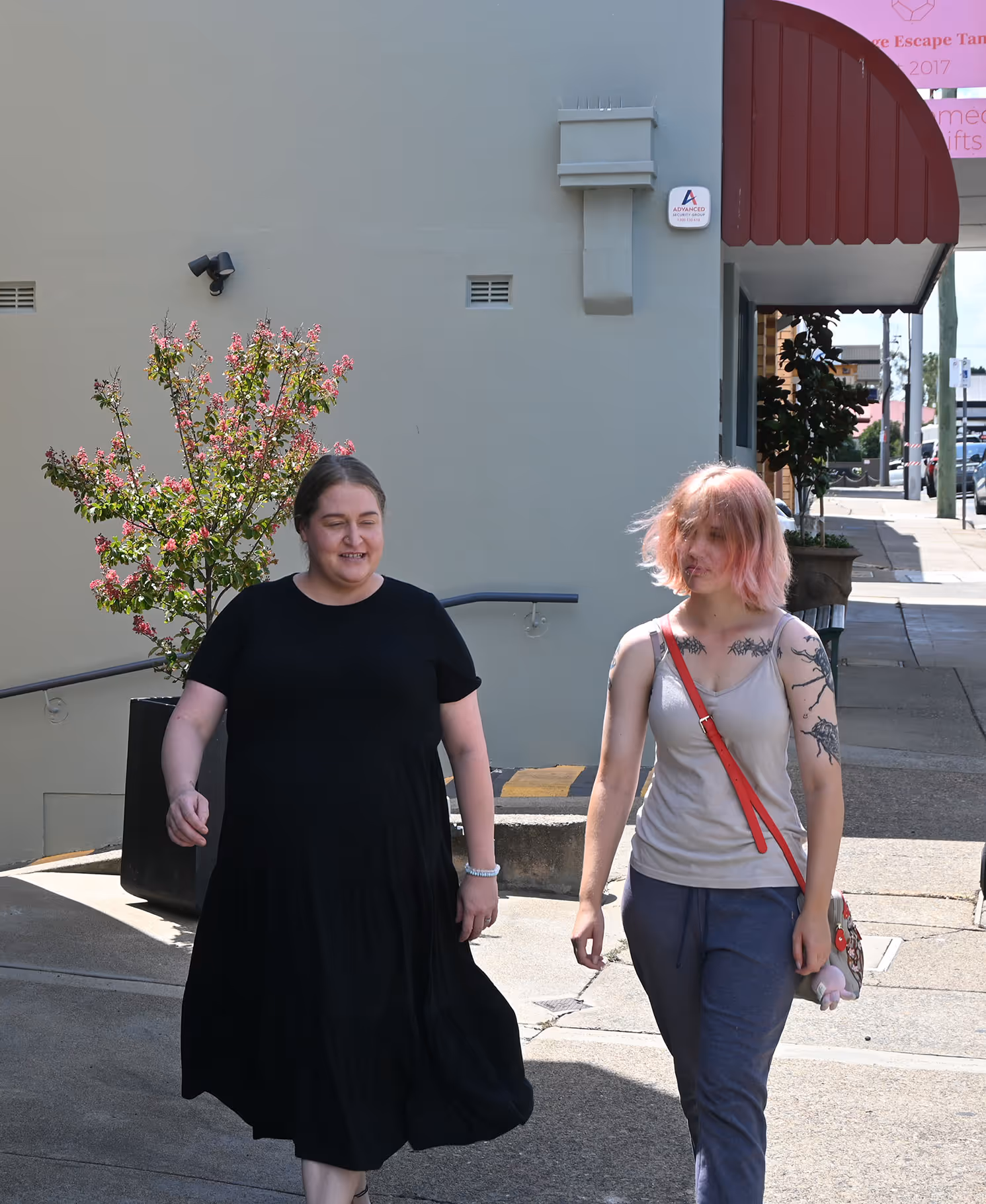 Two women walking on a sunny sidewalk beside a building with a potted flowering tree.