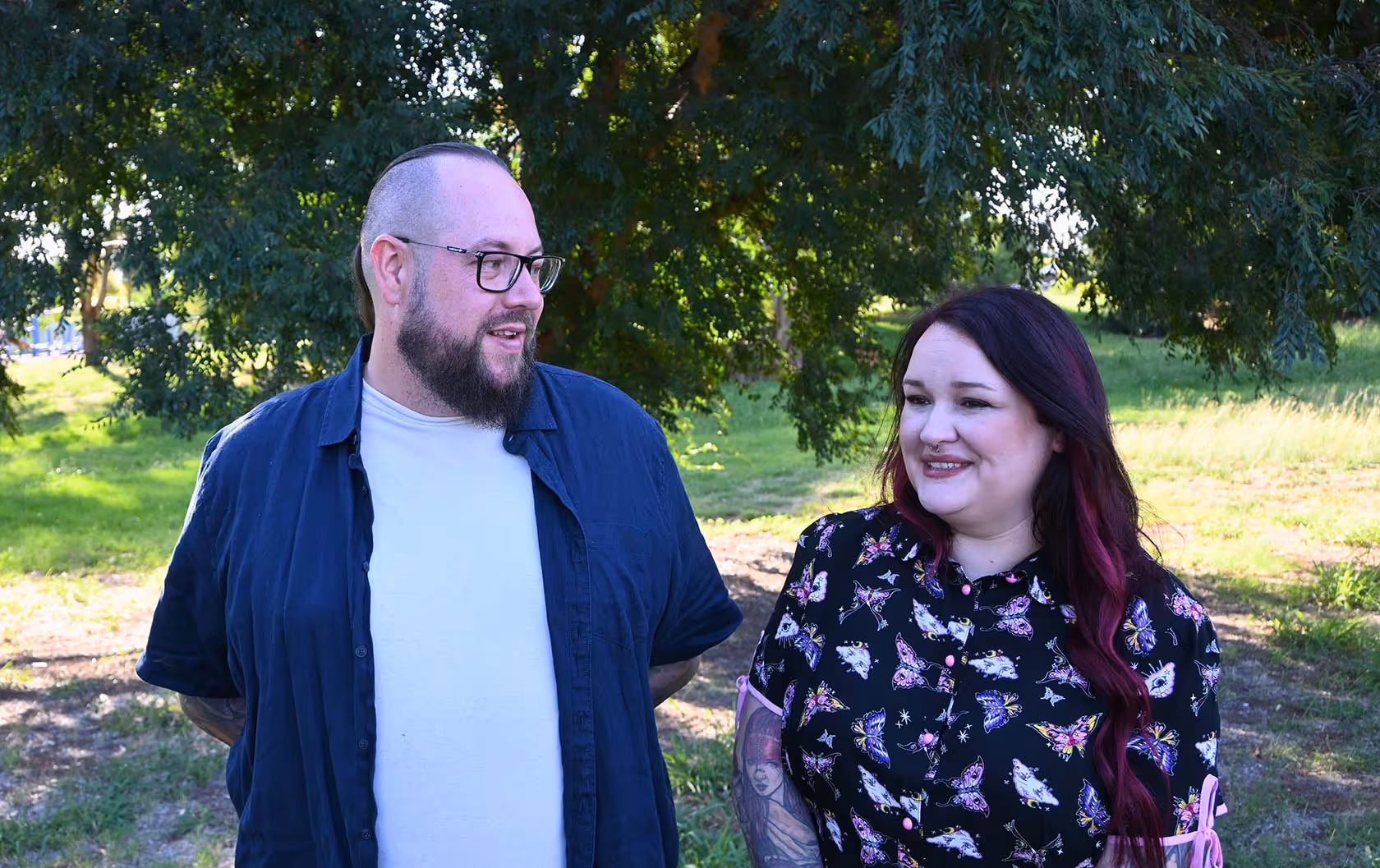 Man with glasses and beard wearing a dark blue shirt standing next to a smiling woman with long dark hair and a butterfly-patterned dress under a tree in a grassy area.